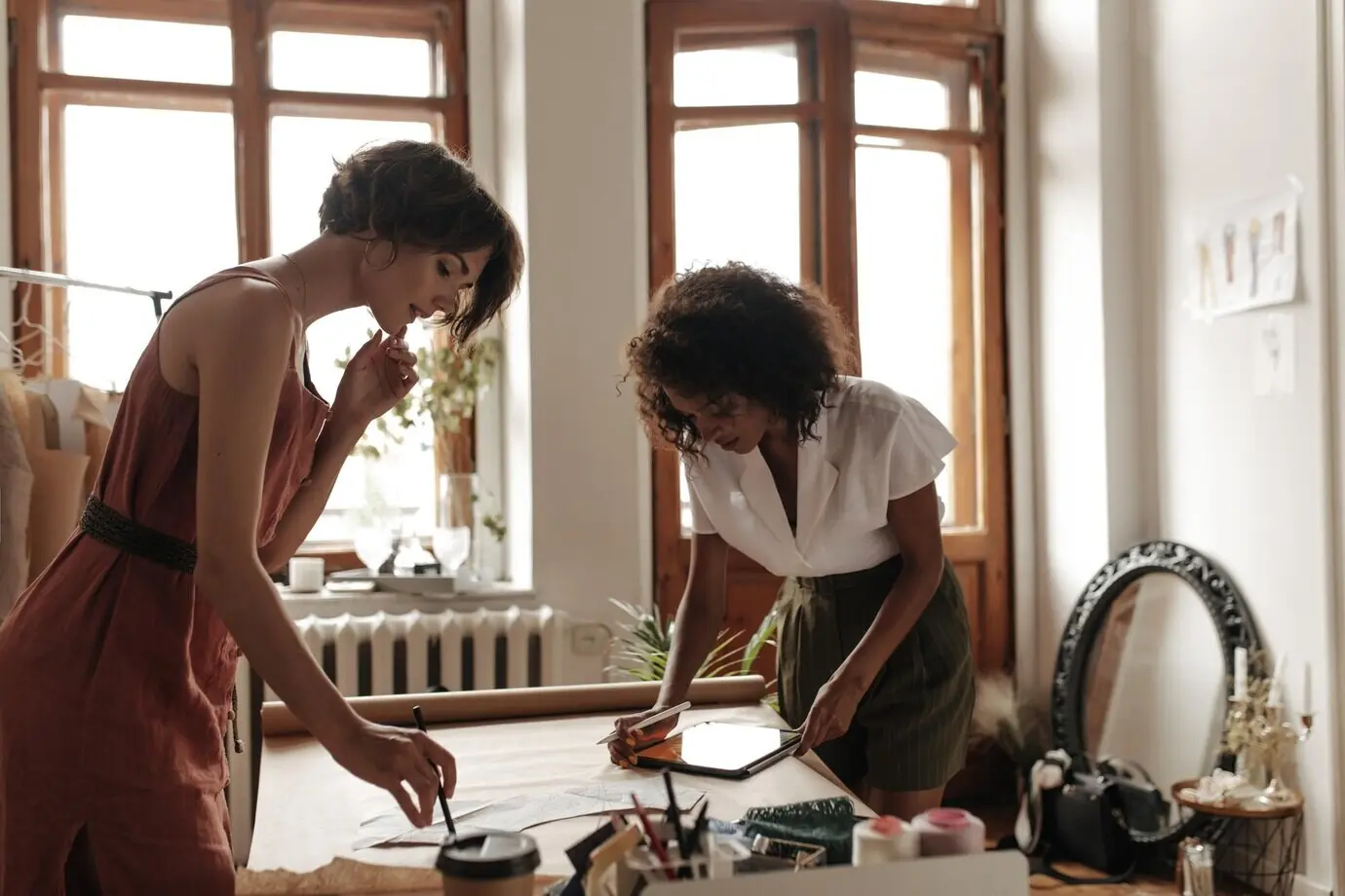Coole junge Frauen in stilvollen Sommeroutfits arbeiten als Modedesignerinnen. Ein Mädchen im roten Kleid und eine Frau in weißer Bluse zeichnen Kleidungsschnittmuster auf einem Blatt Kraftpapier.